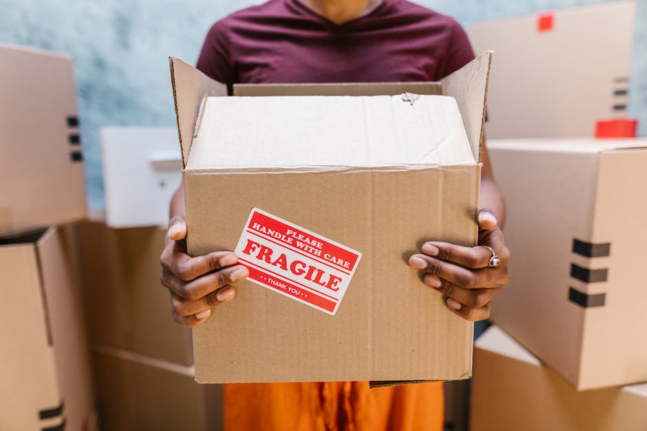 Close-up of a person holding an open cardboard box with a red and white 'Fragile' handling sticker, indicating careful packing for house relocation. The individual's hands are visible, with one hand gripping the side of the box and the other supporting the bottom. The person is wearing a ring on their finger. In the background, there are additional cardboard boxes stacked inside a property, some sealed with black tape and others partially packed. The scene is well-lit, possibly by natural or indoor lighting, emphasizing the careful preparation involved in furniture transport and packing during a house removal. This image illustrates the importance of proper packing and handling in professional removals services, such as those provided by Man and Van Maida Vale, for safe and efficient moving logistics from Little Venice Maida Vale.