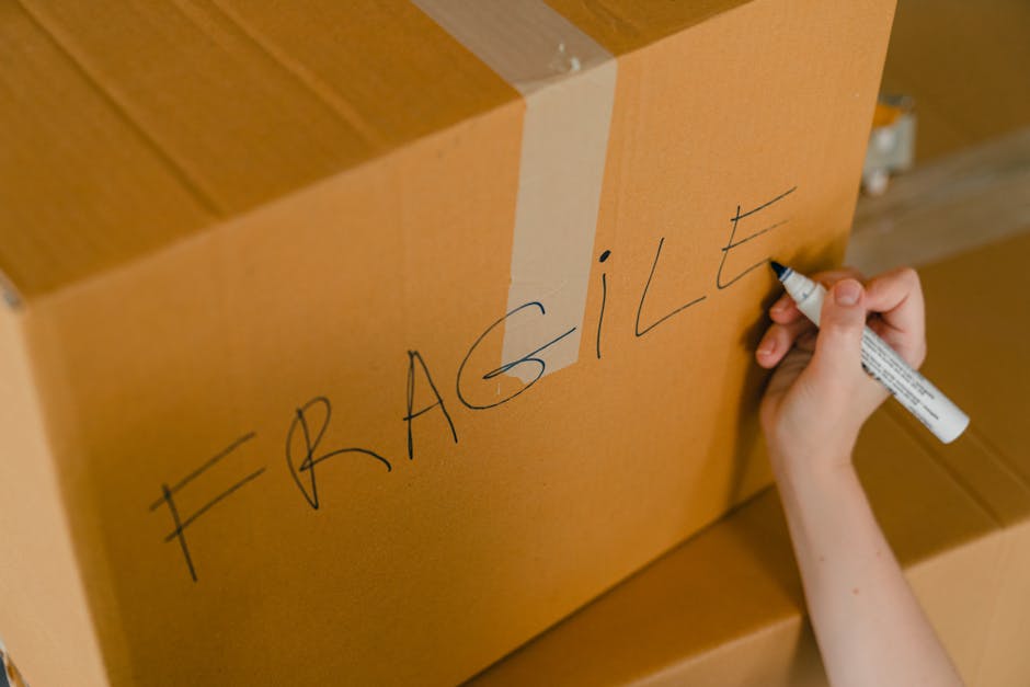 A person's hand is writing the word 'FRAGILE' in large, uppercase letters with a black marker on a brown cardboard box to indicate it contains delicate items. The box is part of a stack of similar boxes, likely used for a house relocation or moving process, placed indoors on a wooden floor. The background shows additional boxes and packaging materials, such as plastic wrap and cardboard padding, used for secure packing of household items. The scene is well-lit, emphasizing the careful handling and packing activities involved in furniture transport and moving services. The image reflects the packaging phase of a professional removal service provided by Man and Van Maida Vale, supporting the safe transportation of goods during home relocations and furniture transport logistics.
