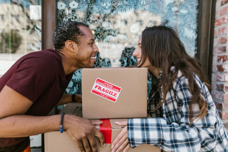 Close-up of a person holding an open cardboard box with a red and white 'Fragile' handling sticker, indicating careful packing for house relocation. The individual's hands are visible, with one hand gripping the side of the box and the other supporting the bottom. The person is wearing a ring on their finger. In the background, there are additional cardboard boxes stacked inside a property, some sealed with black tape and others partially packed. The scene is well-lit, possibly by natural or indoor lighting, emphasizing the careful preparation involved in furniture transport and packing during a house removal. This image illustrates the importance of proper packing and handling in professional removals services, such as those provided by Man and Van Maida Vale, for safe and efficient moving logistics from Little Venice Maida Vale.