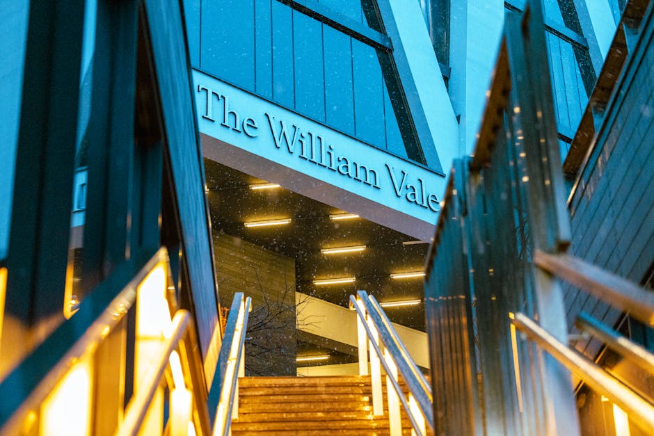 Exterior view of The William Vale building with illuminated signage at dusk, showing a modern structure with dark blue paneling and large glass windows. The entrance features a set of wooden stairs with metal railings leading up to the doorway. On either side of the stairs, there are black metal frames and steps, with some arriving rain visible on the surface. The image captures the heading to a residence or hotel, with the overall environment suggesting a professional home relocation or furniture transport setting. The lighting creates a warm glow around the entrance, highlighting the building's contemporary design, as Man and Van Maida Vale's removals service prepares for packing and loading tasks connected to house moves in the Maida Vale area.