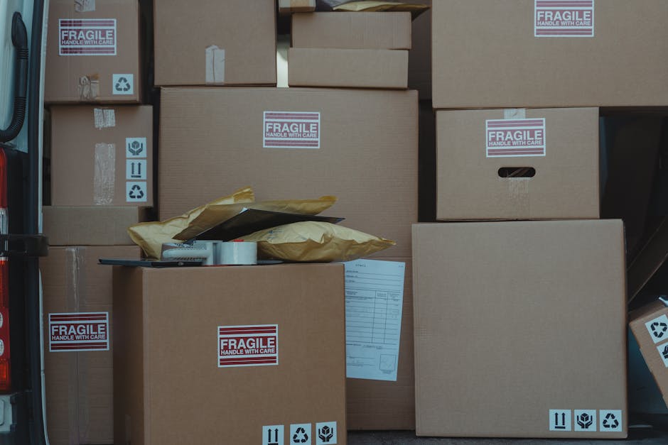 A collection of cardboard boxes, some labeled with 'FRAGILE HANDLE WITH CARE' stickers, are stacked and arranged inside a vehicle, likely a van or moving truck, awaiting or during a home relocation process. The boxes vary in size, and some are sealed with packing tape. A few of the larger boxes are positioned on the pavement or loading area just outside or near the vehicle, ready for transport. Some packaging materials, including brown paper and plastic-wrapped items, are placed on top of the boxes. Recyclable icons and handling instructions are visible on multiple boxes, indicating careful handling during furniture transport and packing for a house move. The scene is well-lit, emphasizing the orderly packing and loading process, which is typical during professional removals by companies like Man and Van Maida Vale, supporting efficient relocation services from start to finish.