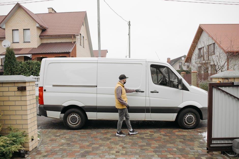 A man wearing a face mask, yellow and gray jacket, and gray pants is standing beside a white commercial van parked on a paved driveway of a residential home, ready to load or unload items during a house removal. The van is positioned next to a brick wall and a metal gate, with the driver’s door open. The background shows a suburban street with two detached houses featuring brickwork, sloped roofs with red tiles, and some window dormers, set against an overcast sky. The scene illustrates the loading process involved in furniture transport and moving logistics, with the van prepared for packing and moving various household belongings as part of a professional removal service near Maida Vale. Man and Van Maida Vale, a company specializing in removals, is visibly engaged in a home relocation activity during this loading process, highlighting safe, efficient furniture transport and packing practices.