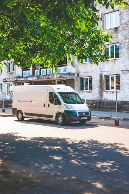 A white cargo van parked on a street in front of a multi-storey residential building, with large windows and a weathered exterior wall. The van is positioned parallel to the pavement, with its side sliding door closed. Overhanging green leaves from a nearby tree cast dappled shadows on the road and the van. The street appears to be part of a residential area in Maida Vale, where Man and Van Maida Vale provides home relocation and furniture transport services. In the background, there is a metal fence along the building's boundary, and the van is ready for a packing and moving process, potentially involving loading furniture and boxes into the vehicle for a house removal. Nearby, an open doorway on the building suggests access for loading or unloading, consistent with professional moving operations.