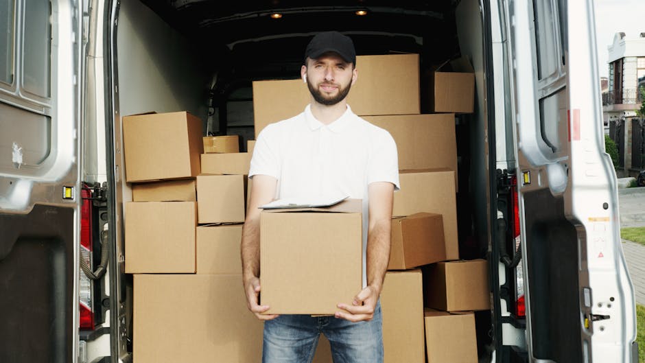 A man wearing a face mask, yellow and gray jacket, and gray pants is standing beside a white commercial van parked on a paved driveway of a residential home, ready to load or unload items during a house removal. The van is positioned next to a brick wall and a metal gate, with the driver’s door open. The background shows a suburban street with two detached houses featuring brickwork, sloped roofs with red tiles, and some window dormers, set against an overcast sky. The scene illustrates the loading process involved in furniture transport and moving logistics, with the van prepared for packing and moving various household belongings as part of a professional removal service near Maida Vale. Man and Van Maida Vale, a company specializing in removals, is visibly engaged in a home relocation activity during this loading process, highlighting safe, efficient furniture transport and packing practices.
