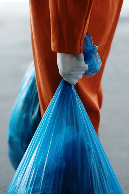 A person wearing a red cardigan and beige trousers is holding a plastic bottle with a white cap, preparing to dispose of it into a designated recycling bin. The recycling container is labeled 'PLASTIC