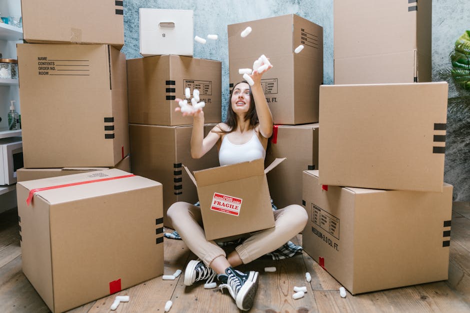 A woman sitting on a wooden floor inside a residential property surrounded by numerous cardboard moving boxes of various sizes, some sealed with red tape and others open. She is wearing a white sleeveless top, beige pants, and black-and-white sneakers, with a joyful expression as she throws packing foam pieces into the air. The boxes are stacked against a light blue textured wall, with a doorway visible behind her leading to another room. Nearby, there are packing materials such as foam and plastic wrap, and part of a white shelf with some bottles on it can be seen to the left. The scene captures the home relocation process, including the packing and lifting activities typical in furniture transport, facilitated by a moving company like Man and Van Maida Vale. Proper lighting highlights the indoor setting, emphasizing the packing materials and the effort involved in preparing for a move.