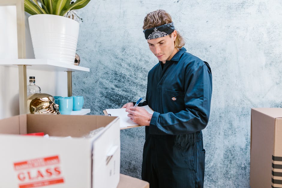 Two removalists from Man and Van Maida Vale inside a well-lit room with arched windows, engaged in a home relocation process. One worker, with dark curly hair and a beard, is holding a medium-sized cardboard box from the top with both hands, standing alongside another worker with light hair, wearing a headband, who is carrying a similar box. Additional cardboard boxes of varying sizes, some labeled, are placed on the wooden floor nearby, ready for transportation. The room has white walls and minimal furniture, including a vintage-style armchair with a wooden frame and green upholstery positioned against the right wall. Natural light filters through the windows, illuminating the scene, which depicts the packing and loading phase of furniture transport during a house removal. This image reflects the logistics involved in professional removals services, emphasizing careful handling and organized packing, aligning with the topic of Maida Vale removals pricing and moving cost guides.