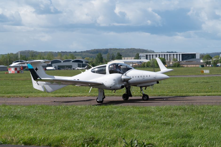 A white aircraft with the word 'Skyborne' on its fuselage is positioned on a tarmac runway at an airport, surrounded by green grass and open fields. The plane has a sleek, modern design with a pointed nose, small wings, and a single engine under each wing. Its landing gear is extended, and the cockpit canopy is closed, with no visible personnel nearby. In the background, there are several hangars and airport buildings, some with curved roofs, and a partly cloudy sky overhead. This scene illustrates the transportation of small aircraft, as part of a house relocation or moving logistics service by Man and Van Maida Vale, with the aircraft possibly being prepared for air transport, reflecting the focus on professional moving and transport solutions for clients.