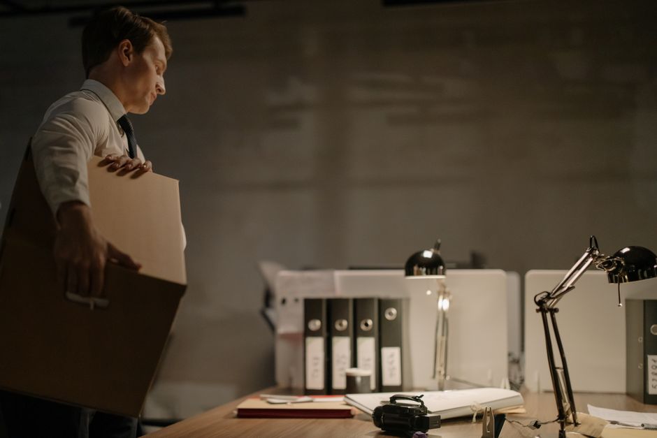 A man with dark hair and a beard, dressed in casual clothing, standing inside a room with white walls and a wooden floor, holding a large cardboard box labelled 'OFFICE' with a smaller label indicating 'MEDIUM'. In the background, there is a metal shelving unit with potted plants and other small items, along with additional packed boxes on the floor, suggesting an ongoing office relocation or packing process. The scene is well-lit with natural light, and the image depicts the process of furniture transport and packing for house removals, consistent with services offered by Man and Van Maida Vale as part of their moving and delivery logistics.