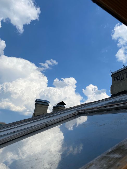 The image shows a view of a bright blue sky with scattered white clouds, seen from inside a property through a partially open roof window. The roof window frame is visible at the top of the image, wit