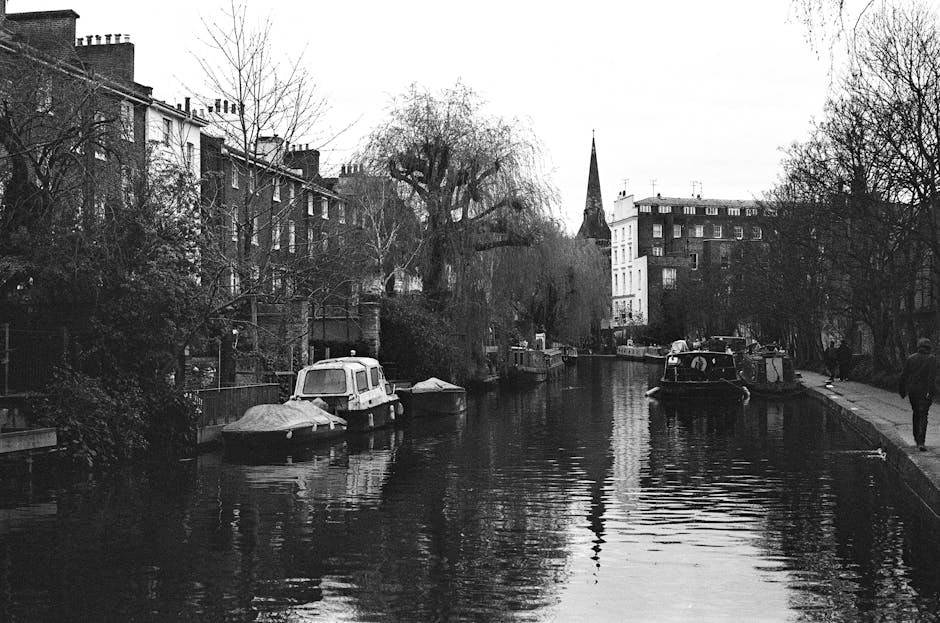 Black and white photograph of a residential canal scene in Maida Vale, London, showing several narrowboats moored along the water's edge, some covered with protective tarps and others exposed. On the right side, a cobbled pathway runs parallel to the canal, with a pedestrian walking along it. To the left, a stone wall and garden area border the canal, with leafless trees extending their branches over the water, indicating a winter setting. Behind the canal, a row of terraced brick houses with chimneys is visible, along with a tall church spire rising in the background. The overall scene reflects a calm, typical London neighbourhood with Victorian architecture, and provides a tranquil setting suitable for house removals or furniture transport services carried out by companies like Man and Van Maida Vale, who specialise in property relocations and moving logistics.