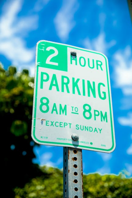 A green and white parking restriction sign mounted on a metal post outdoors, indicating a two-hour parking limit from 8 am to 8 pm, with an exception for Sundays. The sign is positioned above leafy trees and set against a bright blue sky with scattered clouds. The sign includes instructions for parking rules relevant to house removals and property relocations in the Maida Vale area. The sign is slightly tilted, with visible bolts securing it to the post, and the background shows blurred tree foliage, highlighting the urban environment where loading and unloading activities, such as furniture transport and packing during home relocation, may occur near properties serviced by Man and Van Maida Vale.