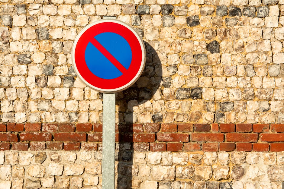 A circular traffic sign with a red border, blue background, and red diagonal line indicating no parking or stopping, mounted on a metal pole against a textured beige and brown brick wall. The sign casts a distinct shadow on the wall, suggesting strong sunlight. The brick wall is composed of irregular stones with mortar, and there is a horizontal red brick line running across the lower part of the wall. The scene is outdoors and appears to be in an urban area, possibly near a property undergoing home relocation or furniture transport. This setting aligns with typical removals and moving services, such as those provided by Man and Van Maida Vale, especially in contexts requiring parking suspensions or parking rule compliance during the loading process.
