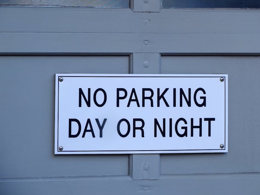 A green and white parking restriction sign mounted on a metal post outdoors, indicating a two-hour parking limit from 8 am to 8 pm, with an exception for Sundays. The sign is positioned above leafy trees and set against a bright blue sky with scattered clouds. The sign includes instructions for parking rules relevant to house removals and property relocations in the Maida Vale area. The sign is slightly tilted, with visible bolts securing it to the post, and the background shows blurred tree foliage, highlighting the urban environment where loading and unloading activities, such as furniture transport and packing during home relocation, may occur near properties serviced by Man and Van Maida Vale.