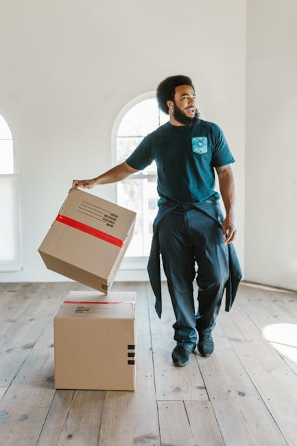 A man with dark, curly hair and a beard, dressed in casual clothing including a blue t-shirt with a logo on the chest and dark pants, is inside a bright, spacious room with white walls and wooden flooring. He is holding a cardboard box wrapped with a red plastic strap, preparing to place it on top of another box on the floor, indicating an ongoing packing or home relocation process. Behind him, there is a large arched window allowing natural light to illuminate the space. The room appears empty aside from the moving boxes, suggesting a furniture transport or packing and moving service. This scene illustrates the manual handling aspect of residential moves handled by companies like Man and Van Maida Vale, which assist with furniture transport and packing logistics during house removals, in line with Westminster Council moving rules and Maida Vale relocation fees.