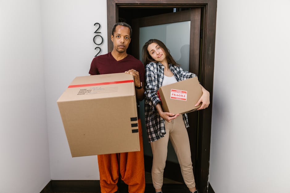 A man and woman stand inside the doorway of an apartment, engaged in a home relocation process as part of a furniture transport and packing and moving service. The man, dressed in a dark red T-shirt and orange trousers, holds a large cardboard box with black markings, while the woman, wearing a plaid shirt over a white top and beige pants, holds a smaller cardboard box labeled 'Fragile' with red and white tape. Both are positioned on the interior side of a black doorframe with a partially open door behind them. The space is well-lit, with white walls and a section of a gray carpeted floor visible. The woman leans her head slightly toward the man, reflecting a casual atmosphere typical of house removals work, and the scene captures the loading process involving multiple boxes, packaging materials, and careful handling as part of a professional moving service offered by Man and Van Maida Vale, in accordance with Westminster Council moving rules and Maida Vale removals fees.